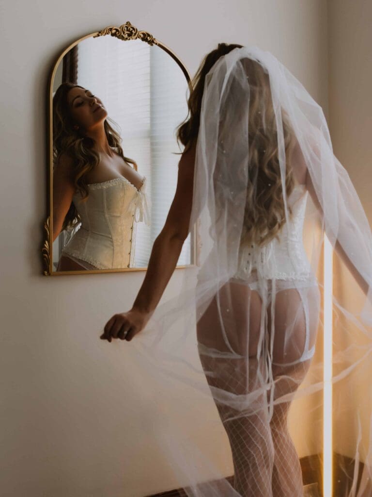 woman posing in front of a mirror wearing bridal lingerie and a veil for her bridal boudoir session