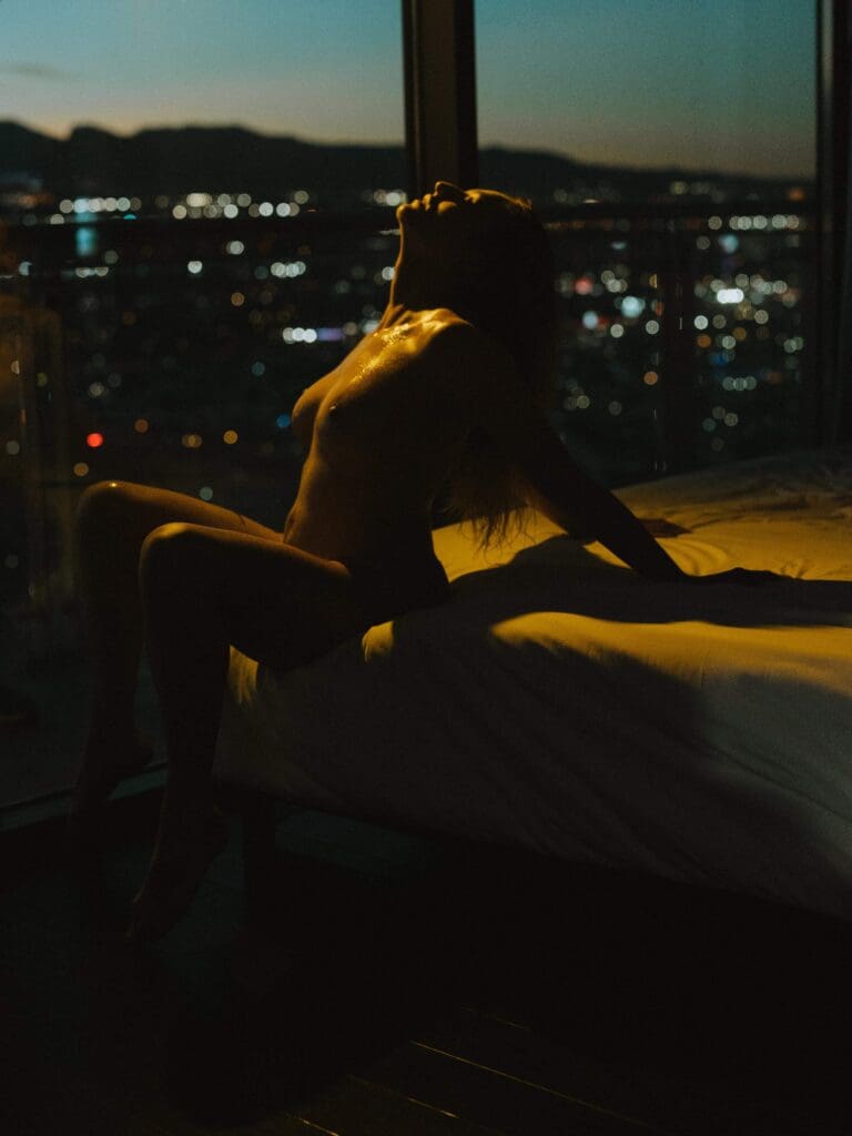 a woman sitting on the edge of the bed leaning her head back during her boudoir photography session, while arching and the beautiful golden lighting accentuates her curvature, with the background of Vegas night lights.
